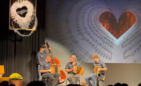 Vier Musiker spielen auf der Bühne Akkordeon, während im Hintergrund ein Herz aus Notenblättern projiziert wird.