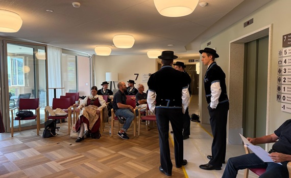 Eine Gruppe von Personen in traditioneller Kleidung steht in einem Raum, während andere auf Stühlen sitzen.