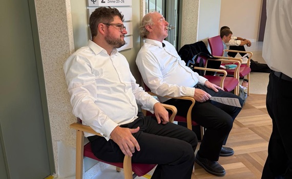 Zwei Männer in weißen Hemden sitzen in einem Flur neben einem Informationsschild.