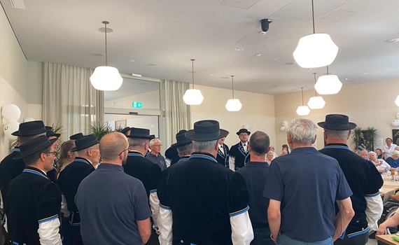 Eine Gruppe von Männern in traditionellen Hüten und Kleidung steht in einem Raum und hört einem Vortrag zu.
