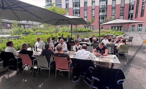 Eine Gruppe von Menschen sitzt an einem Tisch im Freien, umgeben von Pflanzen und Gebäuden.